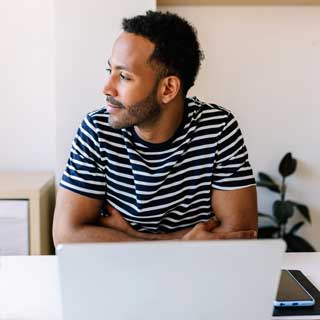 A man in a striped shirt sitting at a desk in front of a laptop, gazing off to the side.