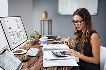 Woman using calculator and computer to review invoices at desk