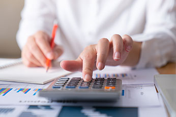 Close-up of person using calculator and writing notes while reviewing financial charts and reports