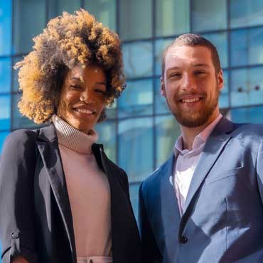 A smiling Black woman and a smiling white man in business casual clothing standing in front of a building looking at the camera after learning about the skills gap.