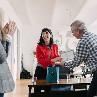 Woman in red jacket receiving certificate and gift bag during office celebration
