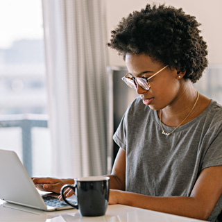 A woman wearing glasses sitting in front of a window at a desk and using a laptop. 