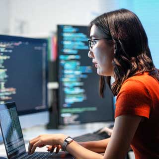 Woman wearing a red shirt and glasses types on a laptop surrounded by cyber security monitoring screens. 