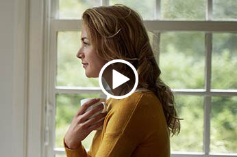 Woman in mustard sweater holding mug, gazing out window at green yard