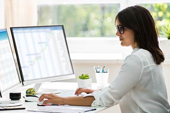 Young professional woman typing on keyboard at dual monitors in sunlit office