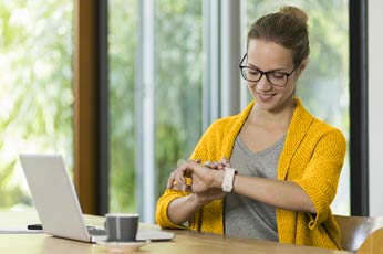Smiling woman checking smartwatch while working at home desk with laptop and coffee