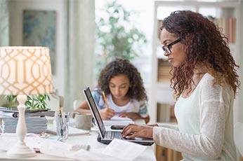Mother working on laptop at home while daughter studies nearby