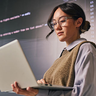 Cyber security professional holds a laptop in front of screens displaying computer code.  