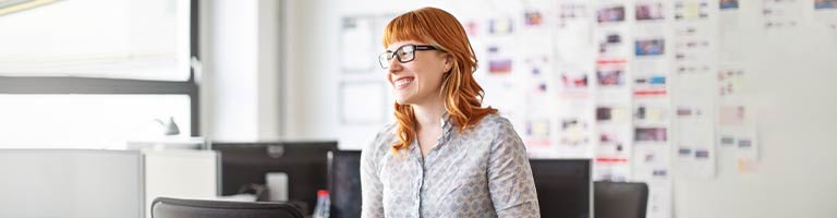 Smiling red-haired woman with glasses standing in bright open office with storyboard