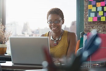 Smiling woman using laptop at windowed desk in colorful creative workspace