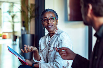 Older woman gesturing and holding tablet while speaking with colleague