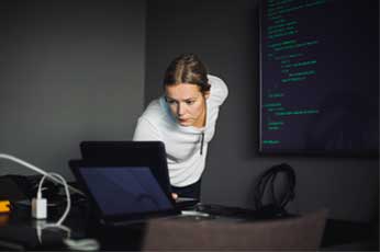 Developer leaning over laptop, reviewing code while monitor displays lines of source code