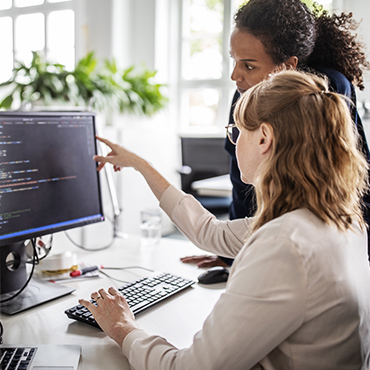 Developer pointing at code on monitor while mentoring colleague in bright open office