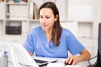 Nurse in blue scrubs using laptop and mouse at clinic desk reviewing patient chart