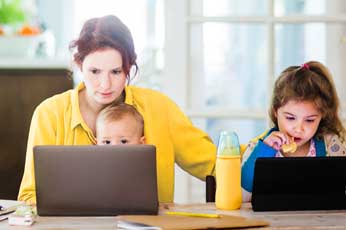 Mother multitasking on laptop while baby watches and daughter uses tablet at kitchen table