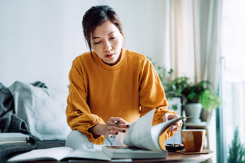 Young woman reviewing documents at coffee table with notebook and mug, bright living room