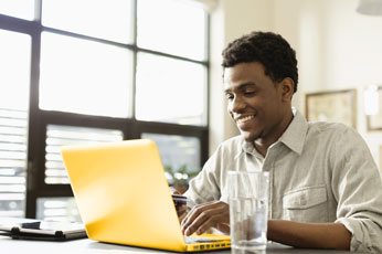 Young man typing on yellow laptop at sunlit home workspace, smiling with smartphone nearby