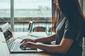 Woman typing on laptop at waterfront cafe table with notebook
