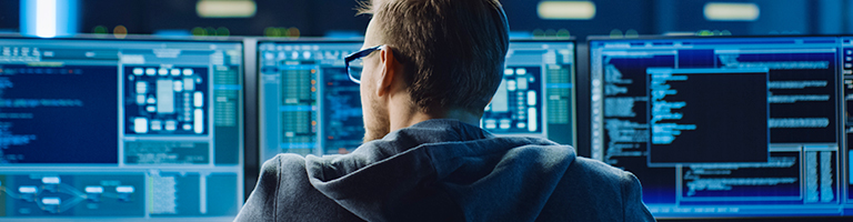 Software engineer wearing glasses and hoodie monitors multiple screens with code
