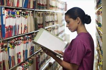Healthcare worker in scrubs reviewing patient file among record shelves.