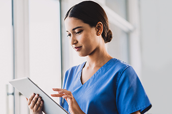 Nurse in blue scrubs reviewing patient information on a tablet beside a bright window
