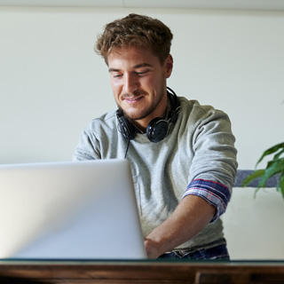 A man working on a laptop, sleeves rolled up, with headphones around his neck.