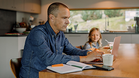 Man studying at his kitchen table with a laptop, notebook, and coffee mug. A young child sits next to him.