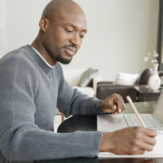 A man in a gray sweater is sitting at a desk, working on a laptop and holding a pencil taking notes. 